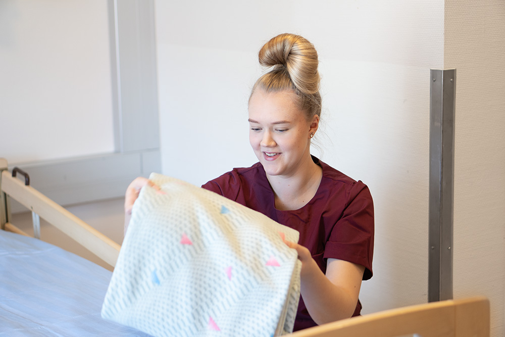 Hoitajanvaatteisiin pukeutunut nuori henkilö viikkaa lakanaa sairaalasänkyyn.